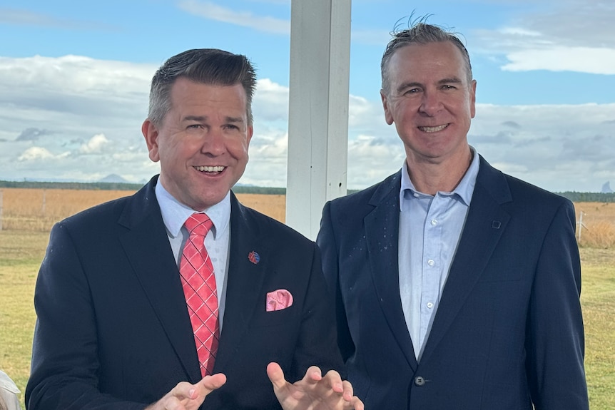 Two smiling men in dark suits at a greenfield site.