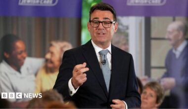 Andy Burnham is wearing a dark-coloured jacket, a white shirt, a tie and glasses. He is speaking to an audience who are not pictured.