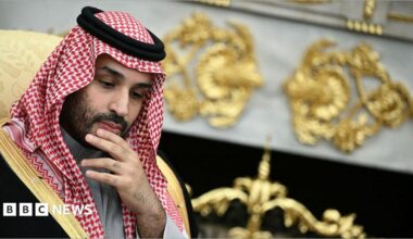 He is rubbing his chin, thinking. He is wearing traditional Arab clothing, with the red and white head dress. He is wearing a black robe with gold edges.
