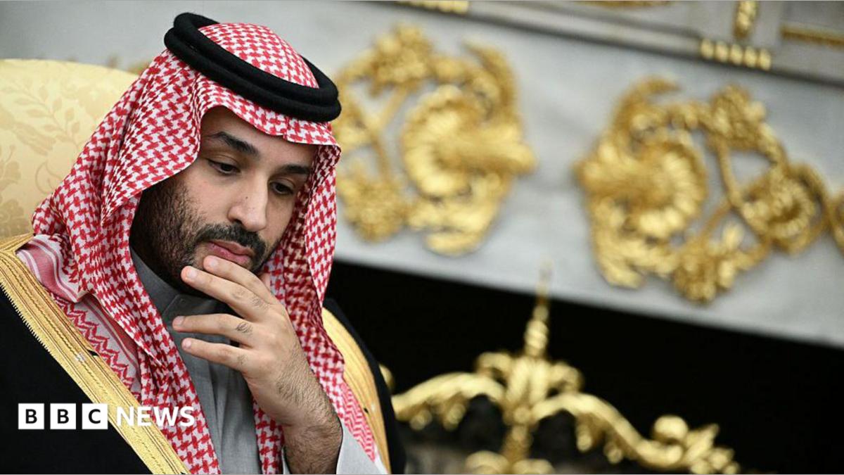 He is rubbing his chin, thinking. He is wearing traditional Arab clothing, with the red and white head dress. He is wearing a black robe with gold edges.