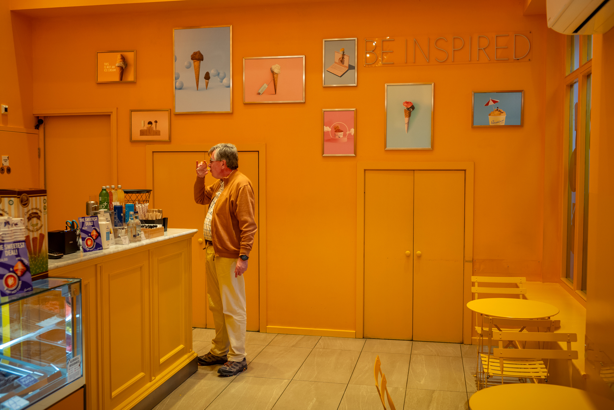 An older man stands by a counter inside a brightly lit ice cream parlor with monochrome yellow-orange walls, artwork of ice cream cones, and matching yellow seating.