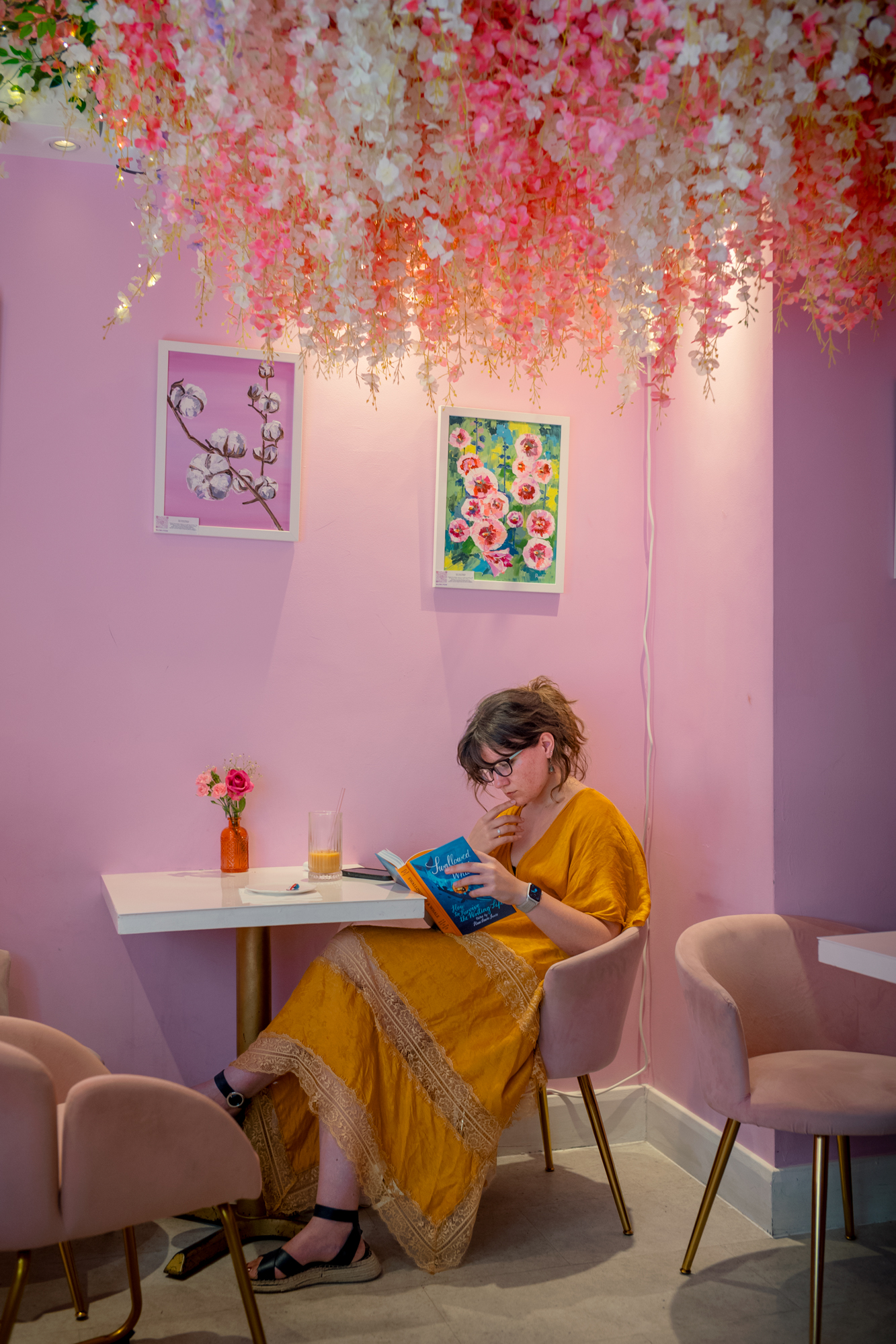 A woman in a mustard-yellow dress sits reading at a small table in a pink café, beneath a canopy of hanging pink and white artificial blossom.