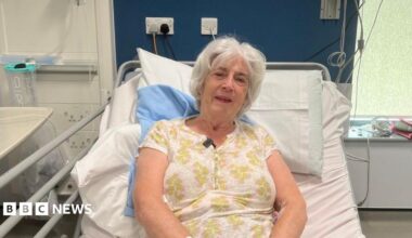 A photo of a woman in a hospital bed, she is wearing a white top with green flowers with some pink on. She is sitting upright and has a little smile. She has white hair.