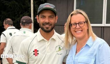 A smiling man dressed in cricket whites is stood next to a smiling woman in a blue shirt at a cricket ground
