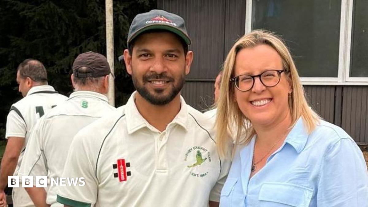 A smiling man dressed in cricket whites is stood next to a smiling woman in a blue shirt at a cricket ground
