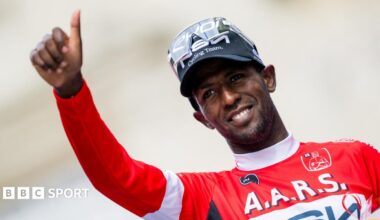 Biniam Girmay, visible from stomach up wearing a long-sleeved green cycling jersey, white cap and sunglasses pushed up on to the top of his head, holds both arms up as he smiles in celebration. He is on a post-race podium, discernible due to the sponsors logos visible on the yellow and green backdrop