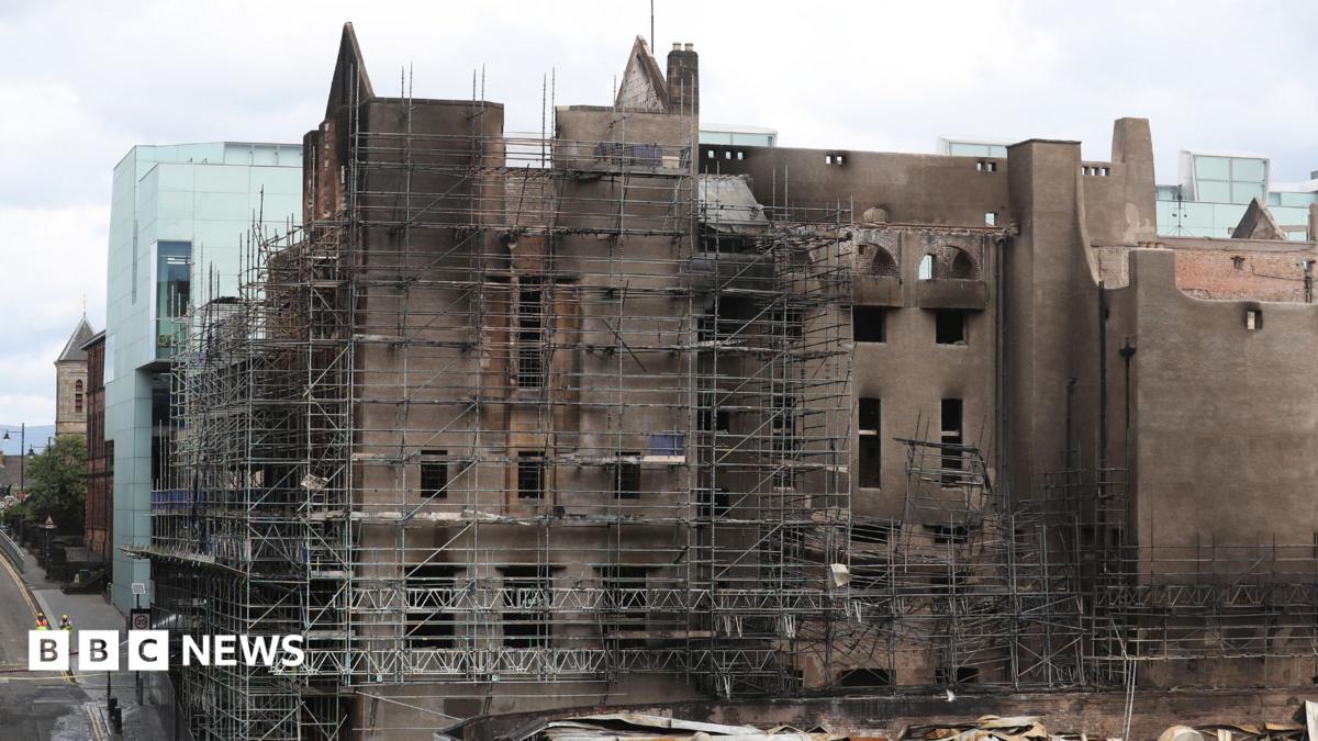 Large historic building undergoing restoration, surrounded by extensive metal scaffolding. The facade shows darkened, fire-damaged walls and empty window openings, while modern glass-sided structures are visible on either side. A wet street runs alongside the building, with two workers in high-visibility clothing visible in the lower left corner under an overcast sky