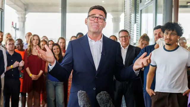 Andy Burnham (C) stands in a room with his arms both raised as he's surrounded by people. He's wearing a dark blue suit and a white shirt