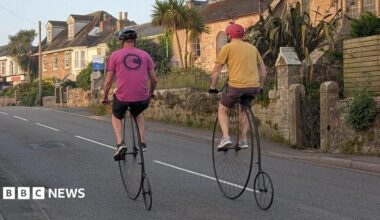 Two riders on penny farthing bicycles cycle along a quiet Cornish street past a stone church.