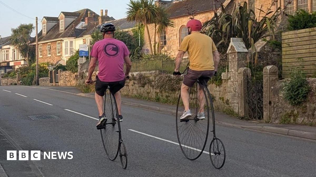 Two riders on penny farthing bicycles cycle along a quiet Cornish street past a stone church.