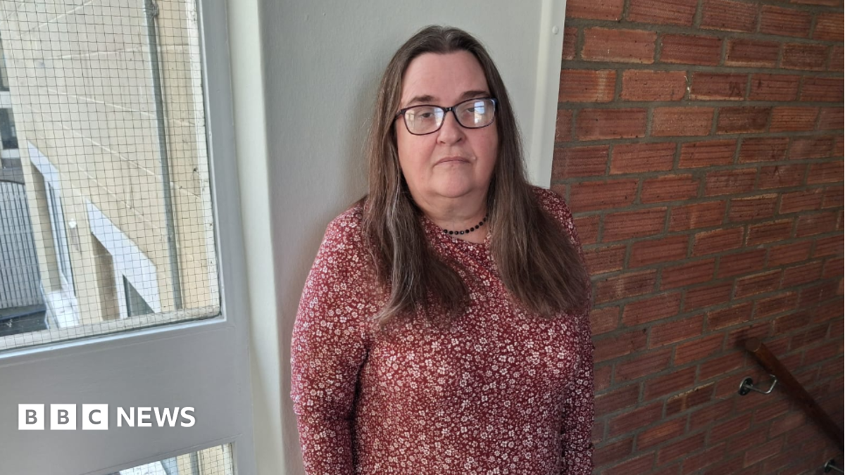 Pearl is standing in the stairwell of a block of flats looking to camera with an unhappy face. She has long brown hair and is wearing black glasses and a red dress with white floral pattern