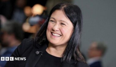 Head and shoulders shot of Nandy, who wears a black t-shirt under a black jacket, and a large red Labour rosette on her lapel. She wears her long black hair loose over her shoulders and is smiling broadly.