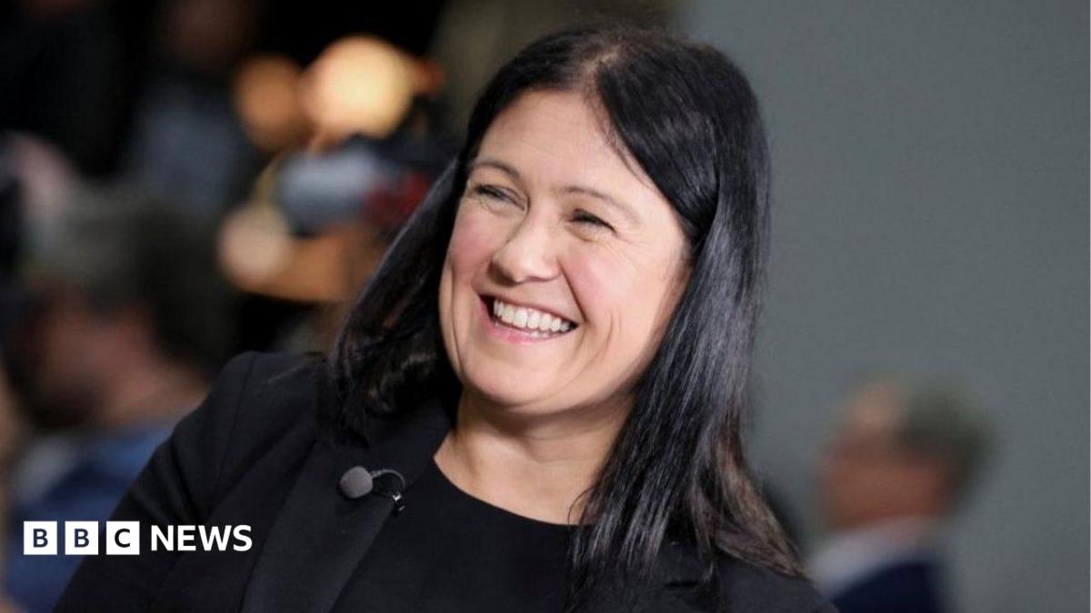 Head and shoulders shot of Nandy, who wears a black t-shirt under a black jacket, and a large red Labour rosette on her lapel. She wears her long black hair loose over her shoulders and is smiling broadly.