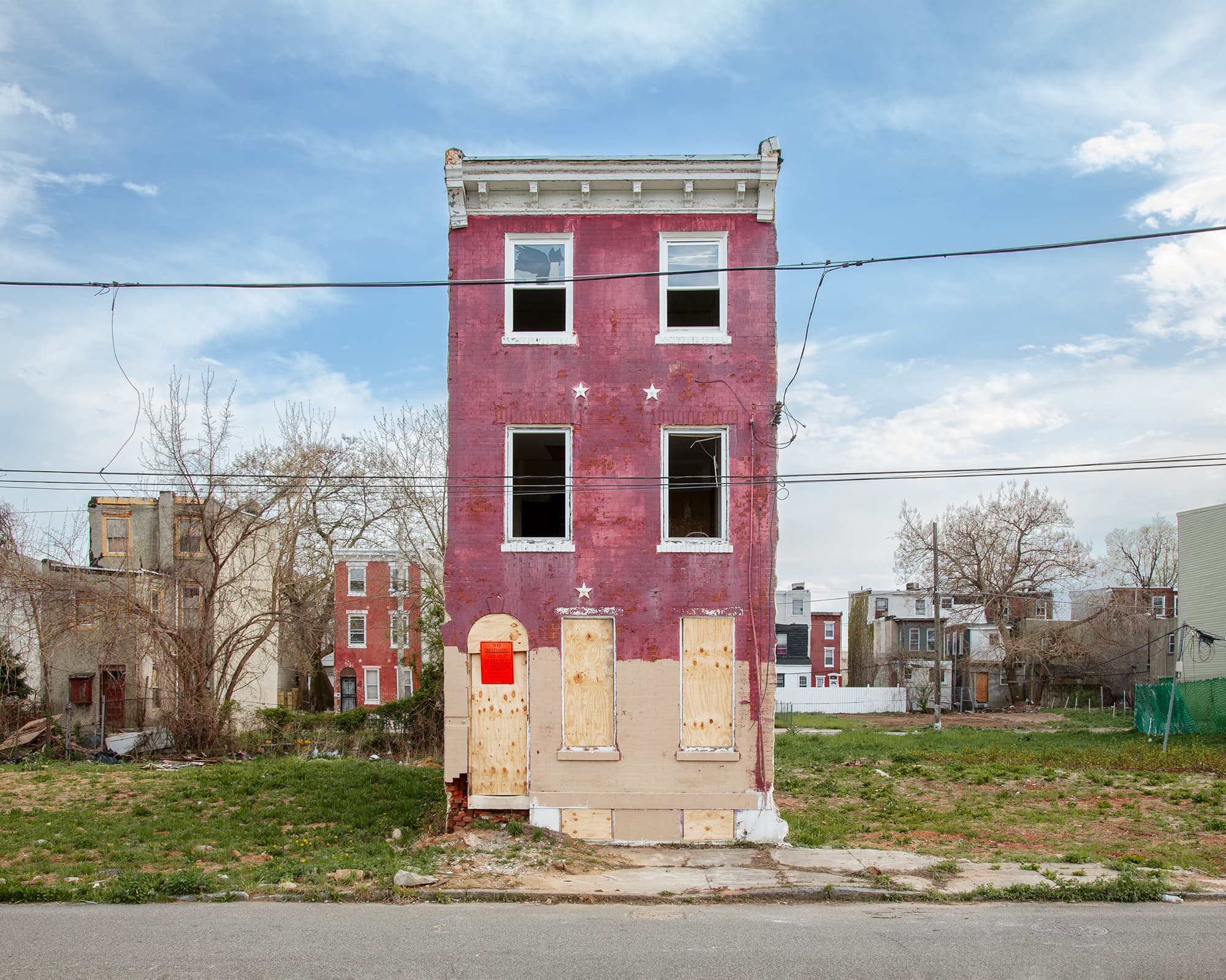 A crumbling, lone three-story pink rowhouse with boarded-up lower windows and open, glassless upper windows sits in an overgrown, vacant lot under a blue sky.