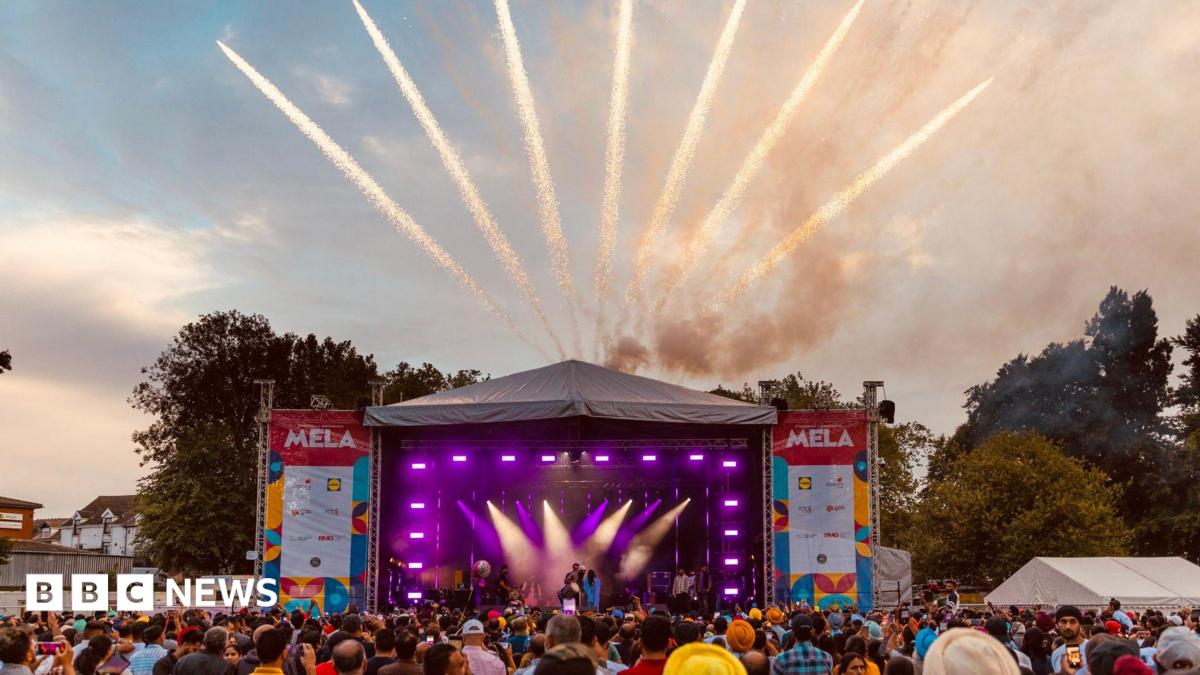 A photo of a stage with purple lighting and crowds of people stood in front of it. Pop-up signs near the stage say 'MELA'. There are sparks of light rising from above the stage.