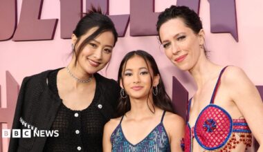 Hottle stands in the centre of the image on a red carpet wearing a blue sparkling dress. Fala Chen wears a black two piece outfit and has her dark hair pinned up. Rebecca Hall wears a bright pink and blue cut-out outfit