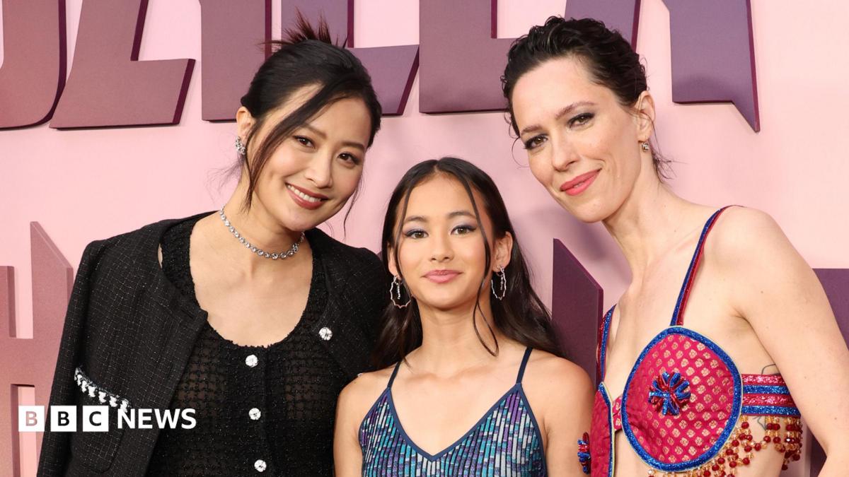 Hottle stands in the centre of the image on a red carpet wearing a blue sparkling dress. Fala Chen wears a black two piece outfit and has her dark hair pinned up. Rebecca Hall wears a bright pink and blue cut-out outfit
