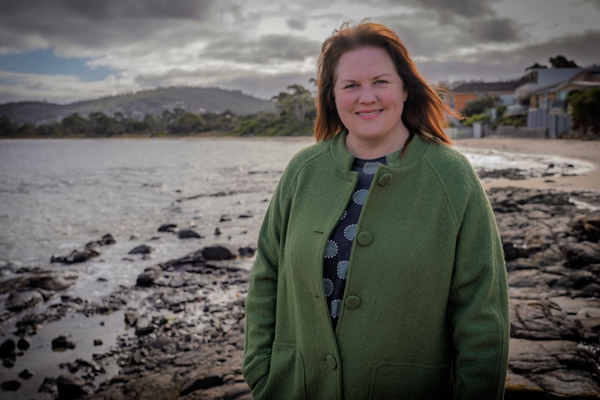 Allison Ritchie wears a green jacket over a blue spotted top and stands on a beach.