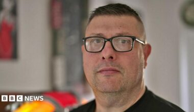 Brian Buckle looks straight towards the camera - he is wearing square glasses and a black jumper. He has short dark hair and dark stubble, and is pictured in a kitchen.