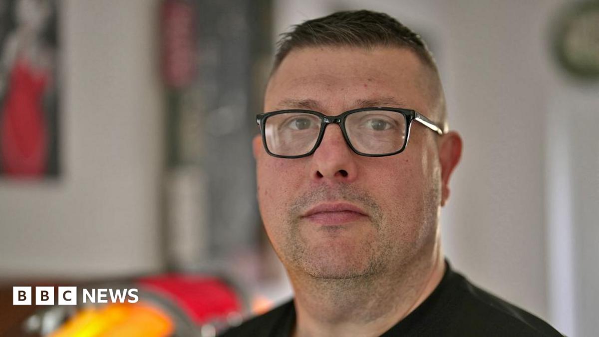Brian Buckle looks straight towards the camera - he is wearing square glasses and a black jumper. He has short dark hair and dark stubble, and is pictured in a kitchen.