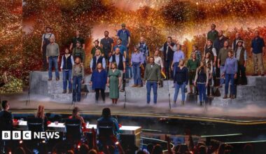 A large group of performers stands on tiered platforms on a brightly lit stage, appearing to sing together as a choir. A conductor stands at the front, guiding the performance. In front of the stage, a panel of judges sits at a table, watching. The background features warm, glowing lights and foliage-like scenery, while an audience in the foreground raises hands and watches the show.