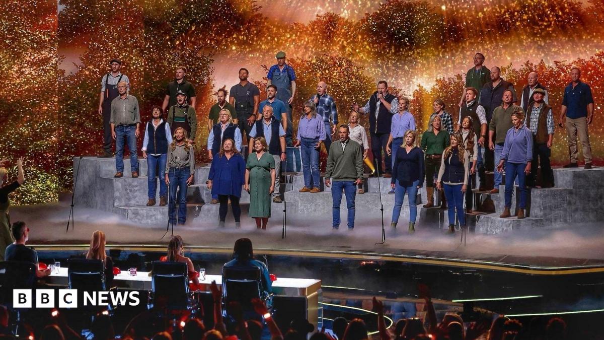 A large group of performers stands on tiered platforms on a brightly lit stage, appearing to sing together as a choir. A conductor stands at the front, guiding the performance. In front of the stage, a panel of judges sits at a table, watching. The background features warm, glowing lights and foliage-like scenery, while an audience in the foreground raises hands and watches the show.