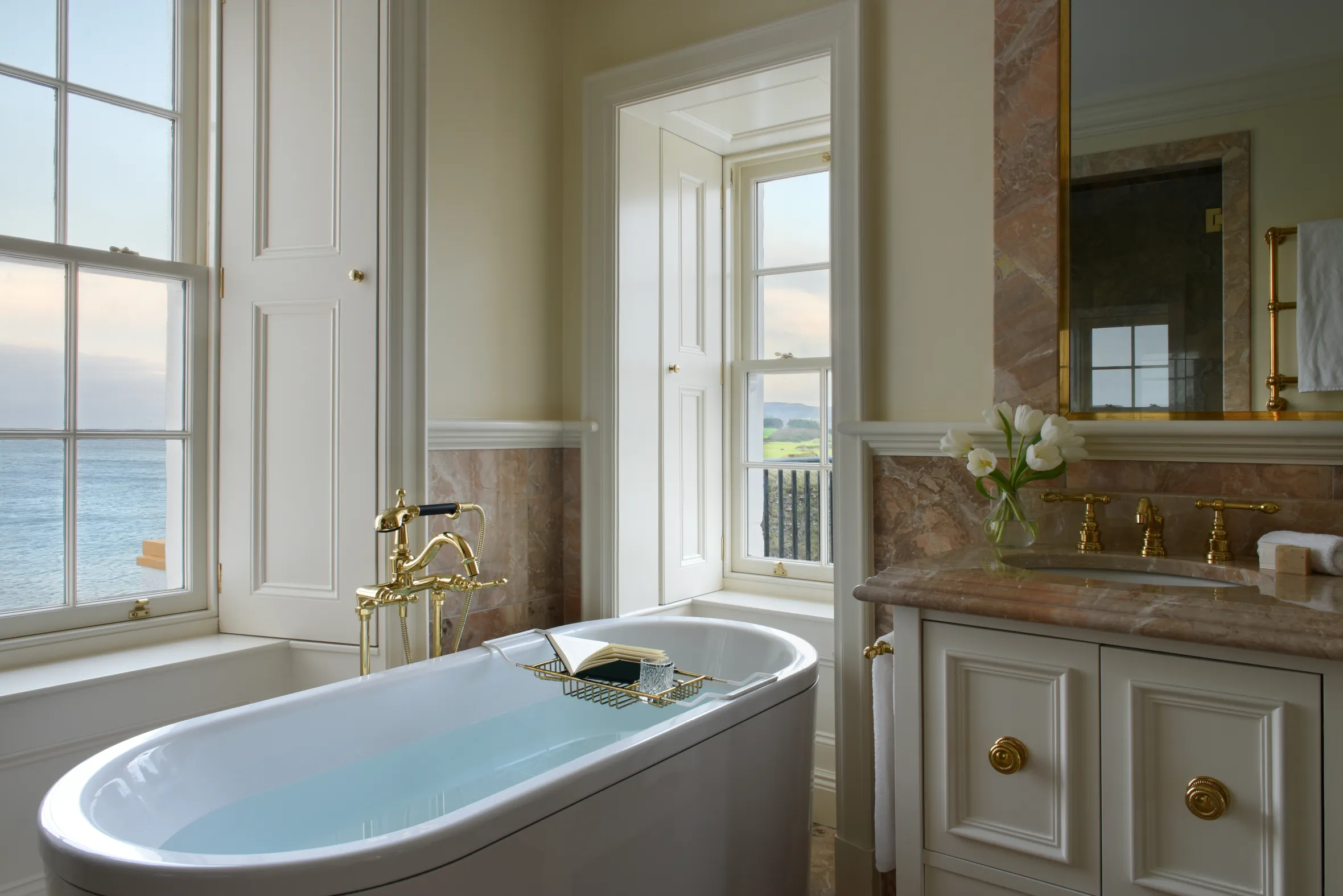 A luxurious white bathtub filled with water, next to a window overlooking the ocean.
