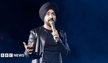 Diljit Dosanjh kicks off his Aura World Tour at BC Place on April 23, 2026, in Vancouver, British Columbia, Canada. He is wearing a black jacket over a black t-shirt and sporting a black turban, as he speaks on a microphone from the stage.