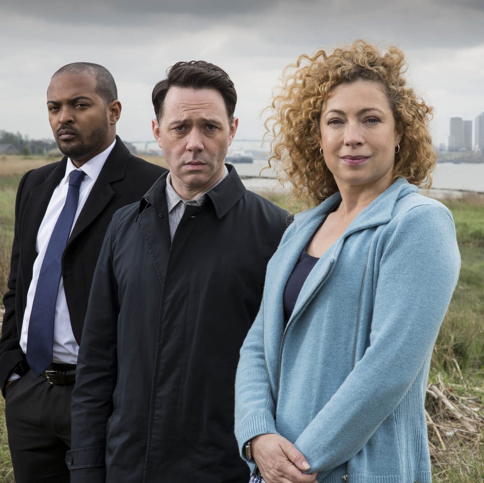 three individuals stand outdoors in an open area with a city skyline in the background three individuals stand outdoors in an open area with a city skyline in the background
