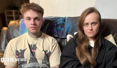 A man and a woman sit together on a sofa facing the camera. The man has a cream T-shirt on with black dogs on it. The woman has long dark brown hair and is wearing a black top