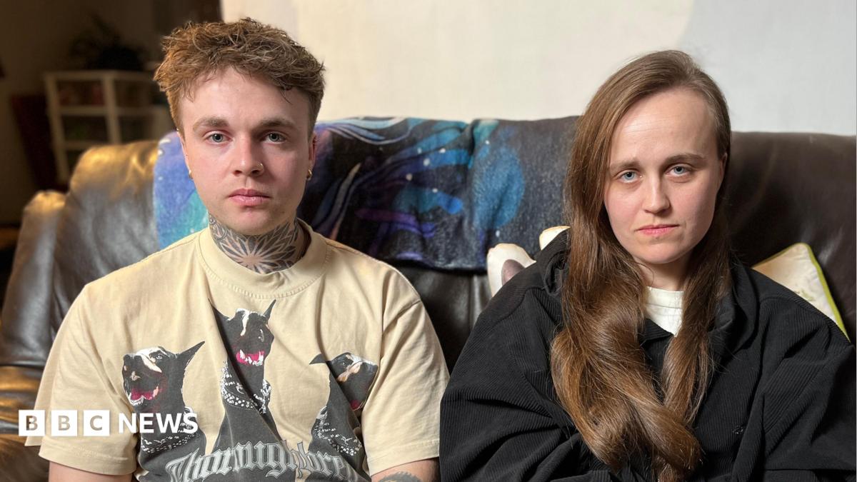 A man and a woman sit together on a sofa facing the camera. The man has a cream T-shirt on with black dogs on it. The woman has long dark brown hair and is wearing a black top
