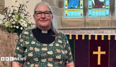 Rev Jane Lee with long grey hair wearing black glasses and a black and white clerical collar with a green cardigan over the top with patterns of sheep and flowers on it. She is standing inside St Wilfrid's Church in Melling near the altar.