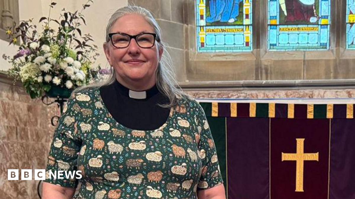 Rev Jane Lee with long grey hair wearing black glasses and a black and white clerical collar with a green cardigan over the top with patterns of sheep and flowers on it. She is standing inside St Wilfrid's Church in Melling near the altar.