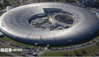 A huge circular silver-roofed building is seen from above, resembling a giant ring. A central open courtyard contains equipment and solar-panel-like arrays. Cars line the perimeter roads, and grass surrounds the complex. The structure looks industrial, modern, and very large.