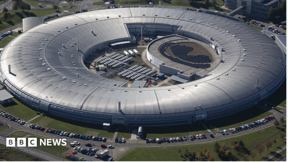 A huge circular silver-roofed building is seen from above, resembling a giant ring. A central open courtyard contains equipment and solar-panel-like arrays. Cars line the perimeter roads, and grass surrounds the complex. The structure looks industrial, modern, and very large.