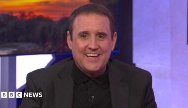 Peter Kay sits on a green sofa, he wears a black knitted polo shirt under a black jacket. He has short dark hair and smiles widely at the camera