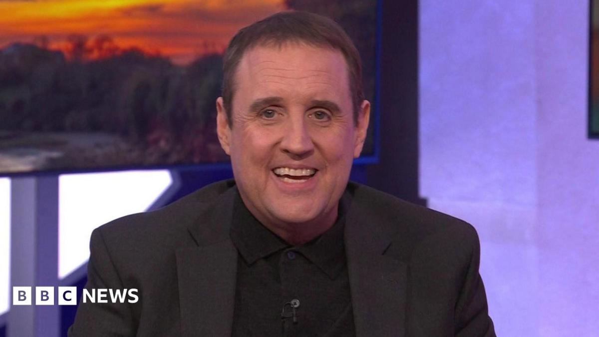 Peter Kay sits on a green sofa, he wears a black knitted polo shirt under a black jacket. He has short dark hair and smiles widely at the camera