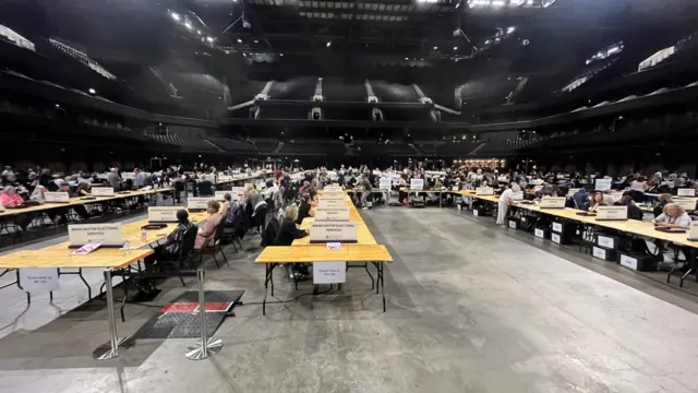 People sit at long rows of tables inside large arena ahead of count