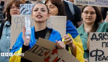 Ukrainians, including a young woman draped in a Ukrainian blue-and-yellow flag, hold placards reading 'Bring him back" during a protest against the dismissal of Defence Minister Mykhailo Fedorov in downtown Kyiv