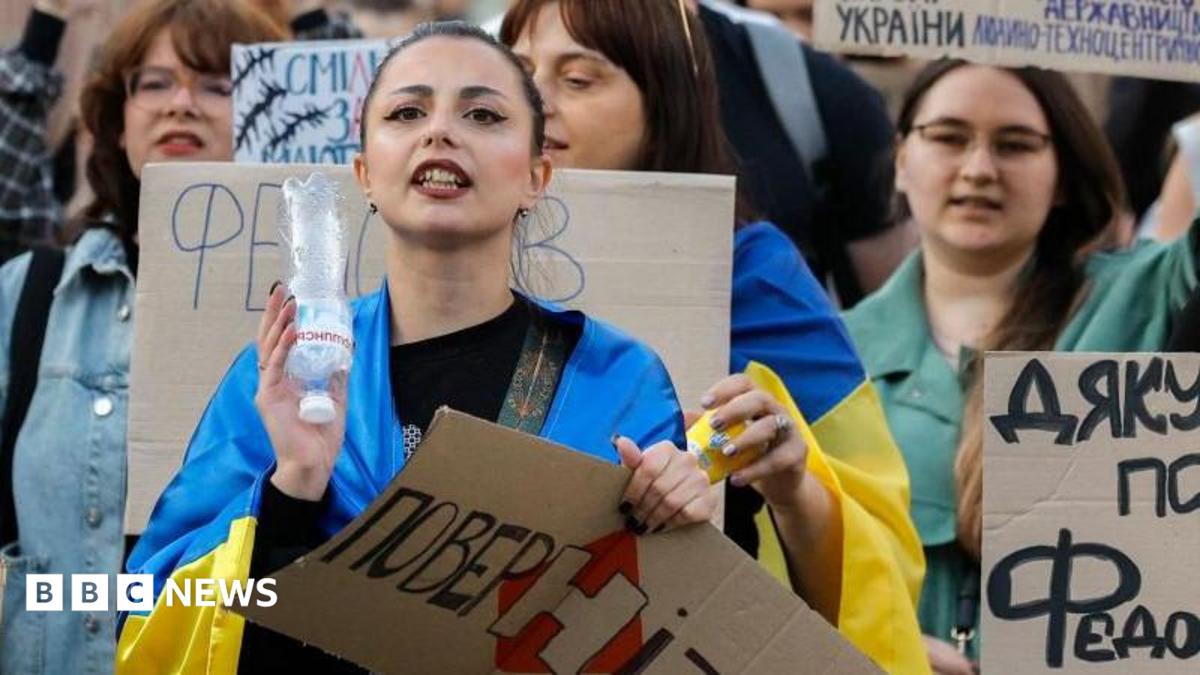 Ukrainians, including a young woman draped in a Ukrainian blue-and-yellow flag, hold placards reading 'Bring him back" during a protest against the dismissal of Defence Minister Mykhailo Fedorov in downtown Kyiv
