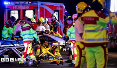 A number of emergency services personnel standing near an ambulance, with a person on a stretcher