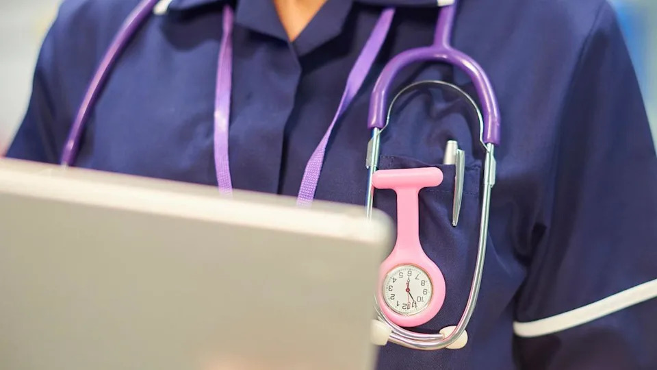 A close-up of a nurse in purple scrubs wearing a stethoscope, using an iPad. Her chest area is visible, but not her face.