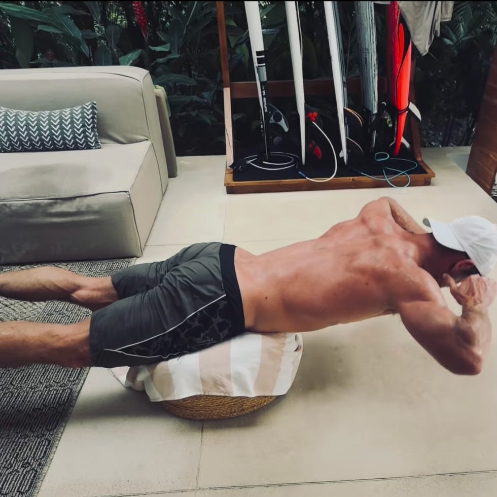man doing back extension exercise on pillow in living room near surfboards.