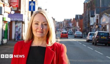Portrait of MP Jo Platt standing on Railway Road, Leigh. She is wearing a red jacket and smiling.