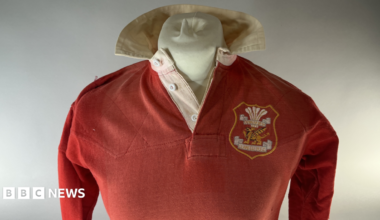 A Llanelli red long sleeved rugby jersey from the historic 1972 win over the All Blacks worn by Tommy David which is being sold at auction. It is shown on a mannequin bust with white background for auction catalogue. It has a white collar, with the club crest on the right hand side of the jersey.