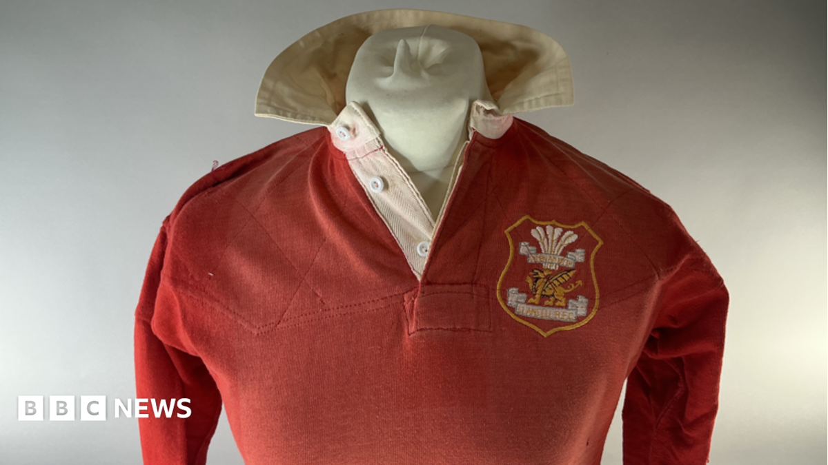 A Llanelli red long sleeved rugby jersey from the historic 1972 win over the All Blacks worn by Tommy David which is being sold at auction. It is shown on a mannequin bust with white background for auction catalogue. It has a white collar, with the club crest on the right hand side of the jersey.
