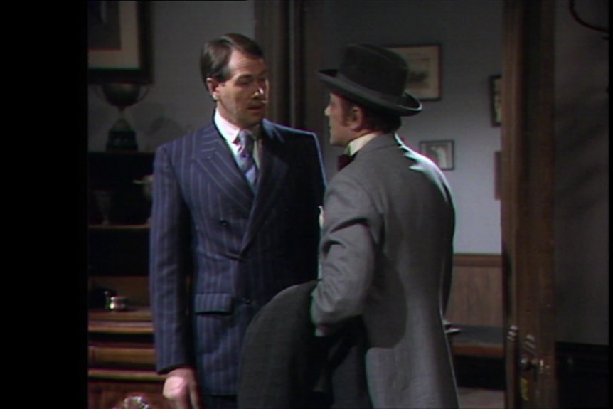 A dark-haired man in a blue and white pinstriped suit stands looking at a man in a grey jacket and black hat.
