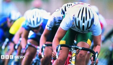 Three male cyclists with white tops and green shorts on a bike racing behind each other. All are wearing white cycling helmets.