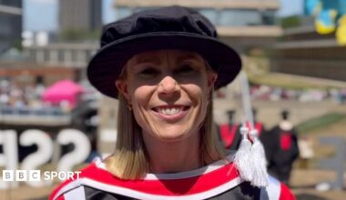 Beth Barrett-Wild in a doctoral-level academic dress, including a black Tudor bonnet hat. She is smiling at the camera and the background is blurred.