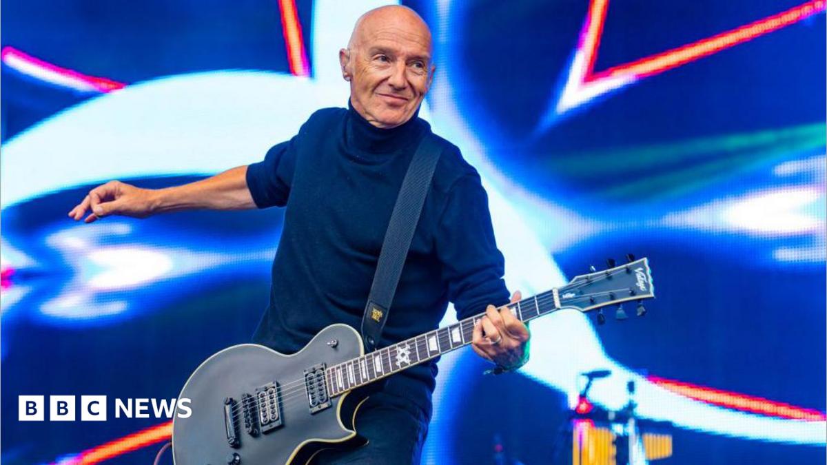a bald man waring a black top is playing an electric guitar on a stage with a light show behind him